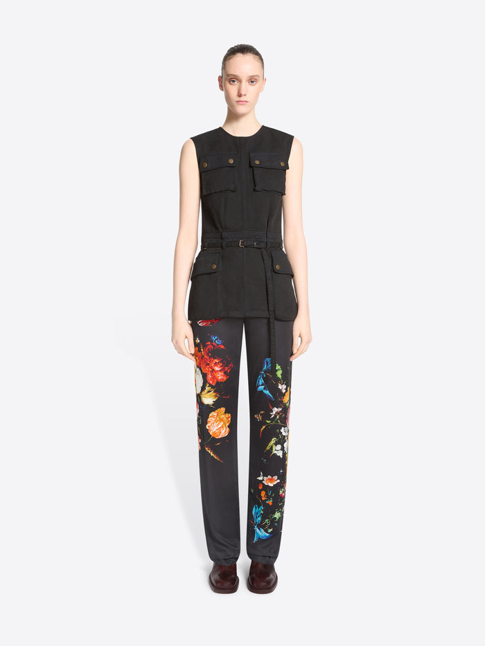 A model stands against a light gray background wearing a sleeveless black utility-style top with flap pockets and black pants featuring a colorful floral print.