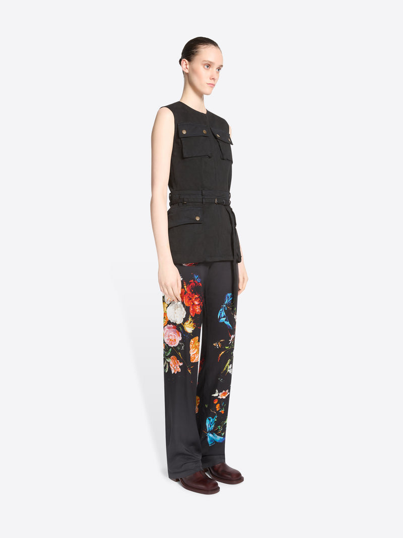 A model stands against a light gray background wearing a sleeveless black utility-style top with flap pockets and black pants featuring a colorful floral print.