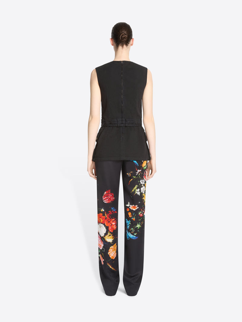 Back view of a model standing against a light gray background wearing a sleeveless black utility-style top with flap pockets and black pants featuring a colorful floral print.