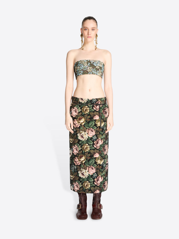 A model stands against a light gray background wearing a matching strapless bandeau top and high-waisted maxi skirt set with a dense floral print, paired with dark brown ankle boots.