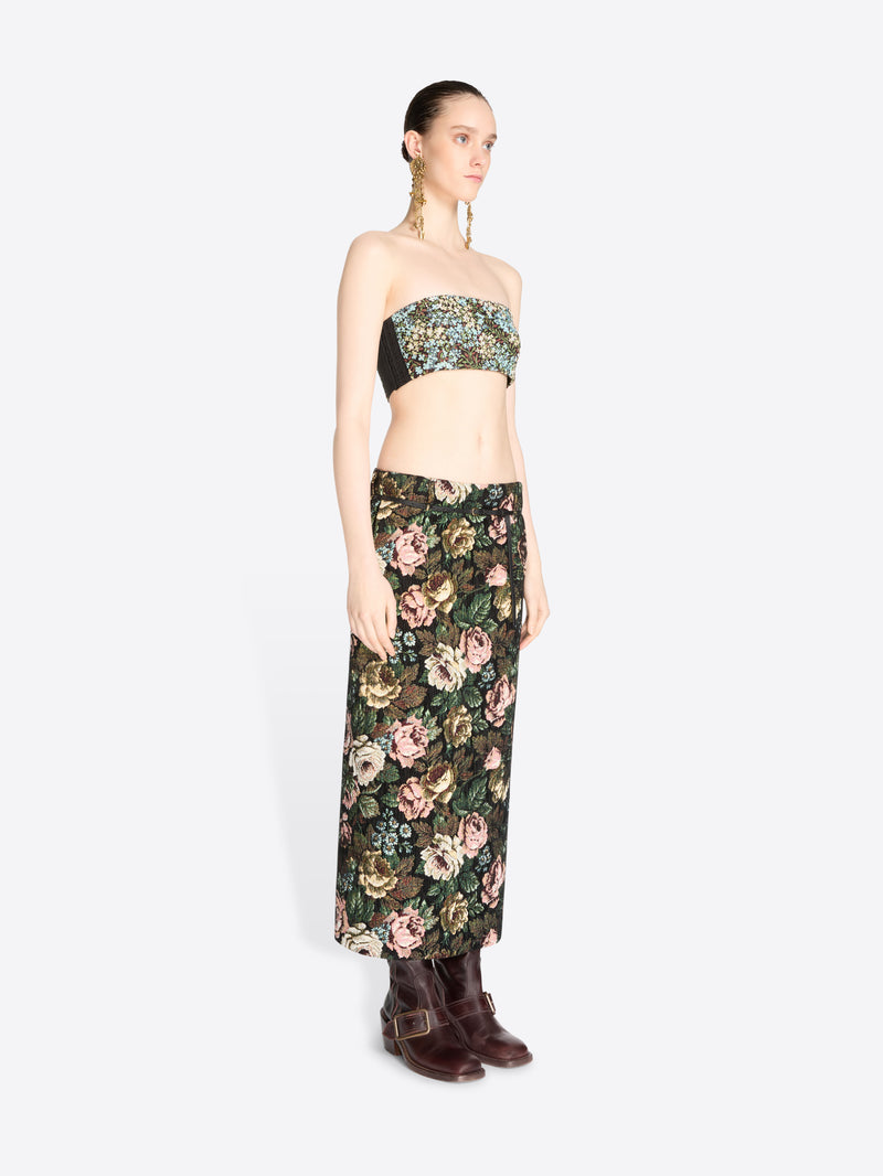 A model stands against a light gray background wearing a matching strapless bandeau top and high-waisted maxi skirt set with a dense floral print, paired with dark brown ankle boots.