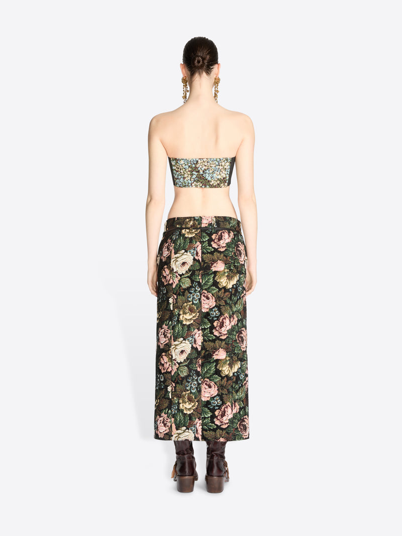 Back view of a model standing against a light gray background wearing a matching strapless bandeau top and high-waisted maxi skirt set with a dense floral print, paired with dark brown ankle boots.