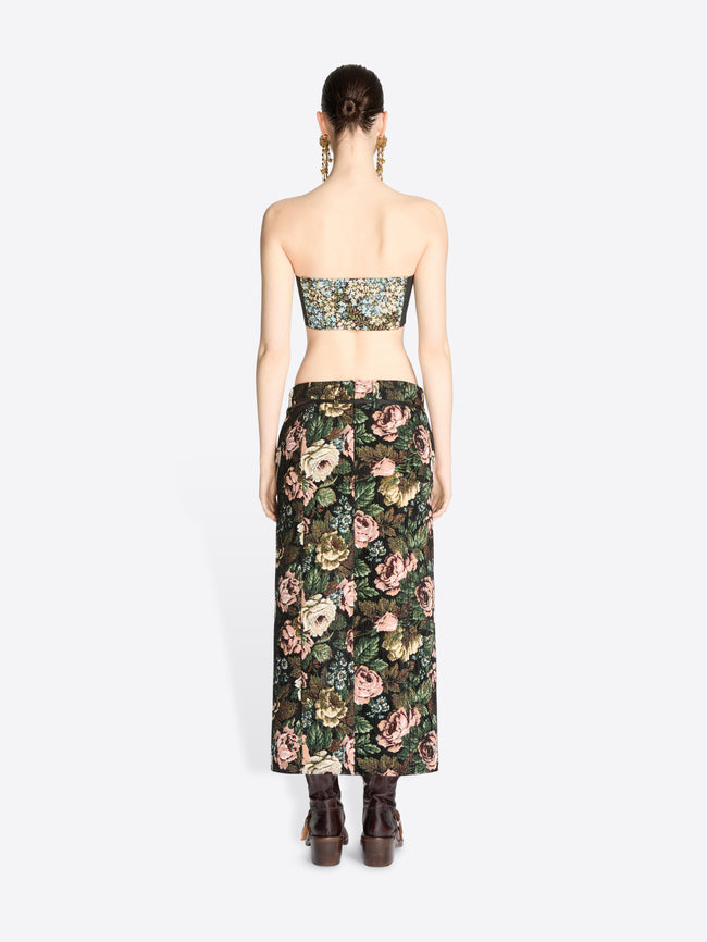 Back view of a model standing against a light gray background wearing a matching strapless bandeau top and high-waisted maxi skirt set with a dense floral print, paired with dark brown ankle boots.