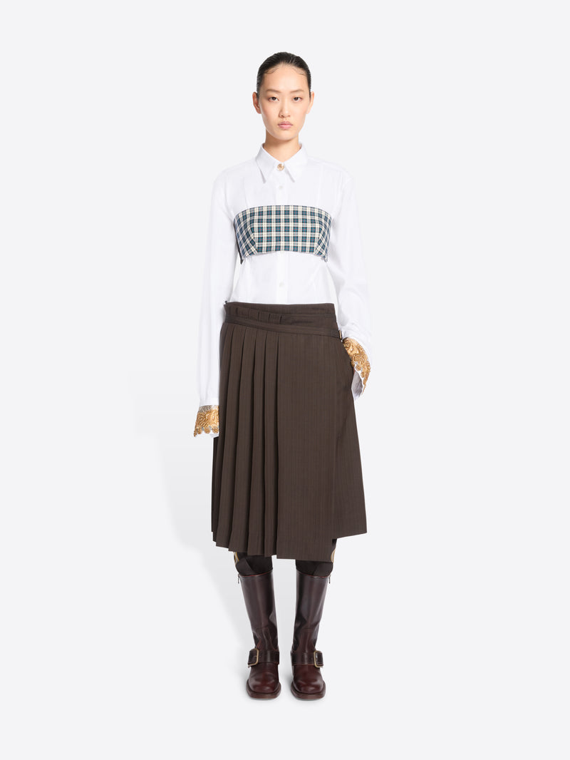 A model stands against a light gray background wearing a white long-sleeve collared shirt, with a green and white  plaid chest top, a dark brown pleated midi skirt, and dark brown knee-high boots.