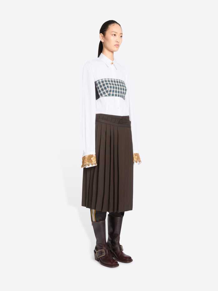 A model stands against a light gray background wearing a white long-sleeve collared shirt, with a green and white  plaid chest top, a dark brown pleated midi skirt, and dark brown knee-high boots.