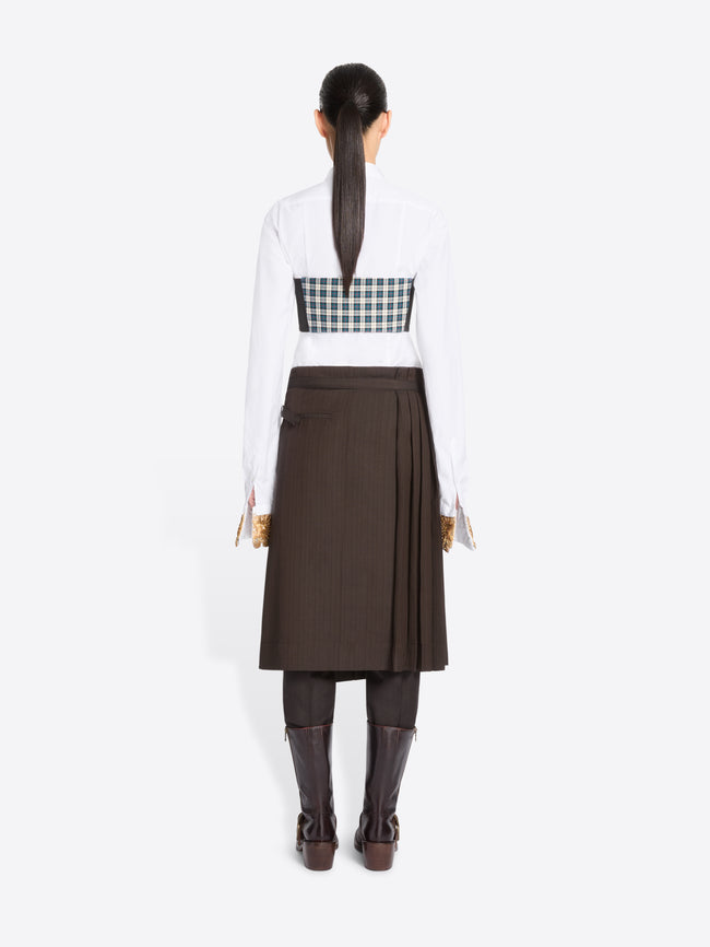 Back view of a model standing against a light gray background wearing a white long-sleeve collared shirt, with a green and white  plaid chest top, a dark brown pleated midi skirt, and dark brown knee-high boots.