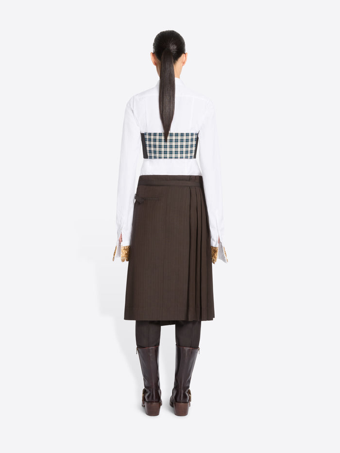 Back view of a model standing against a light gray background wearing a white long-sleeve collared shirt, with a green and white  plaid chest top, a dark brown pleated midi skirt, and dark brown knee-high boots.