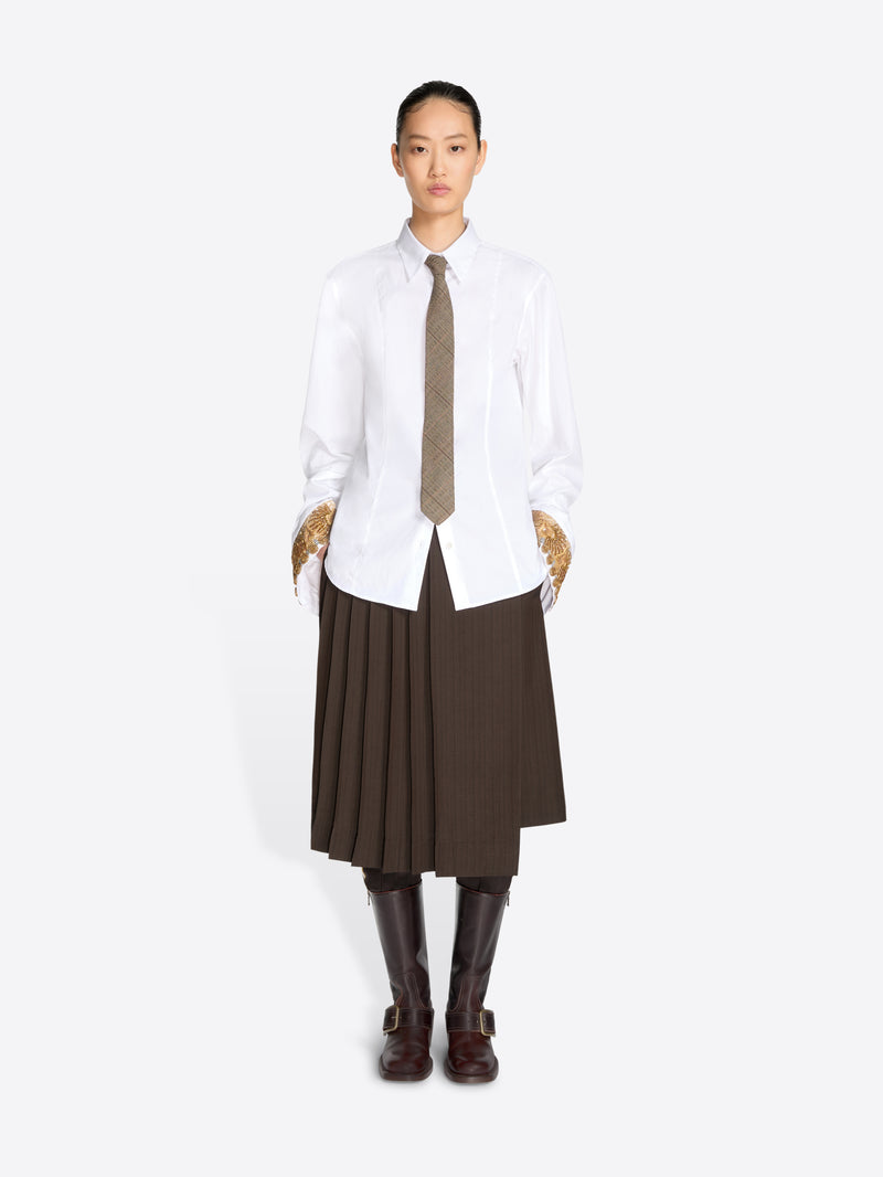 A model stands against a light gray background wearing a white long-sleeve button-up shirt, a brown pleated midi skirt, a brown tie, and dark brown knee-high boots.