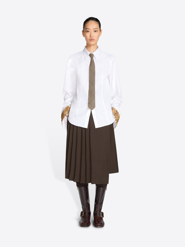 A model stands against a light gray background wearing a white long-sleeve button-up shirt, a brown pleated midi skirt, a brown tie, and dark brown knee-high boots.