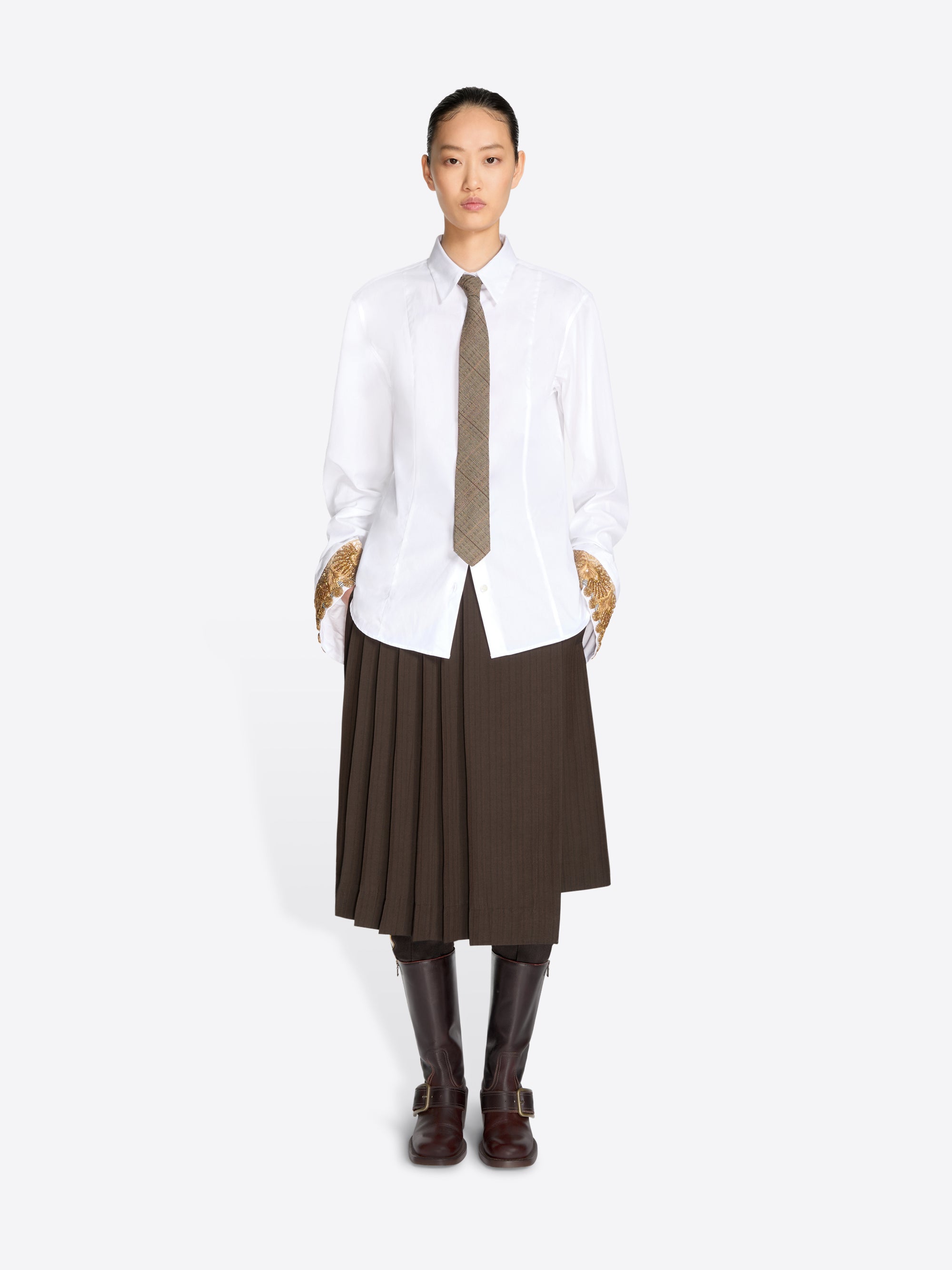 A model stands against a light gray background wearing a white long-sleeve button-up shirt, a brown pleated midi skirt, a brown tie, and dark brown knee-high boots.