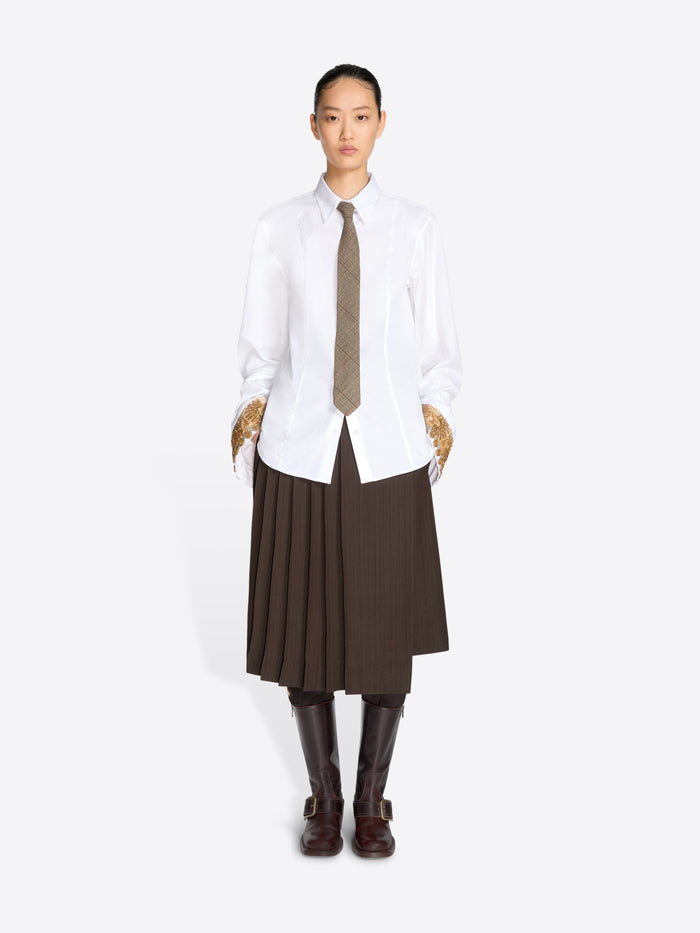 A model stands against a light gray background wearing a white long-sleeve button-up shirt, a brown pleated midi skirt, a brown tie, and dark brown knee-high boots.