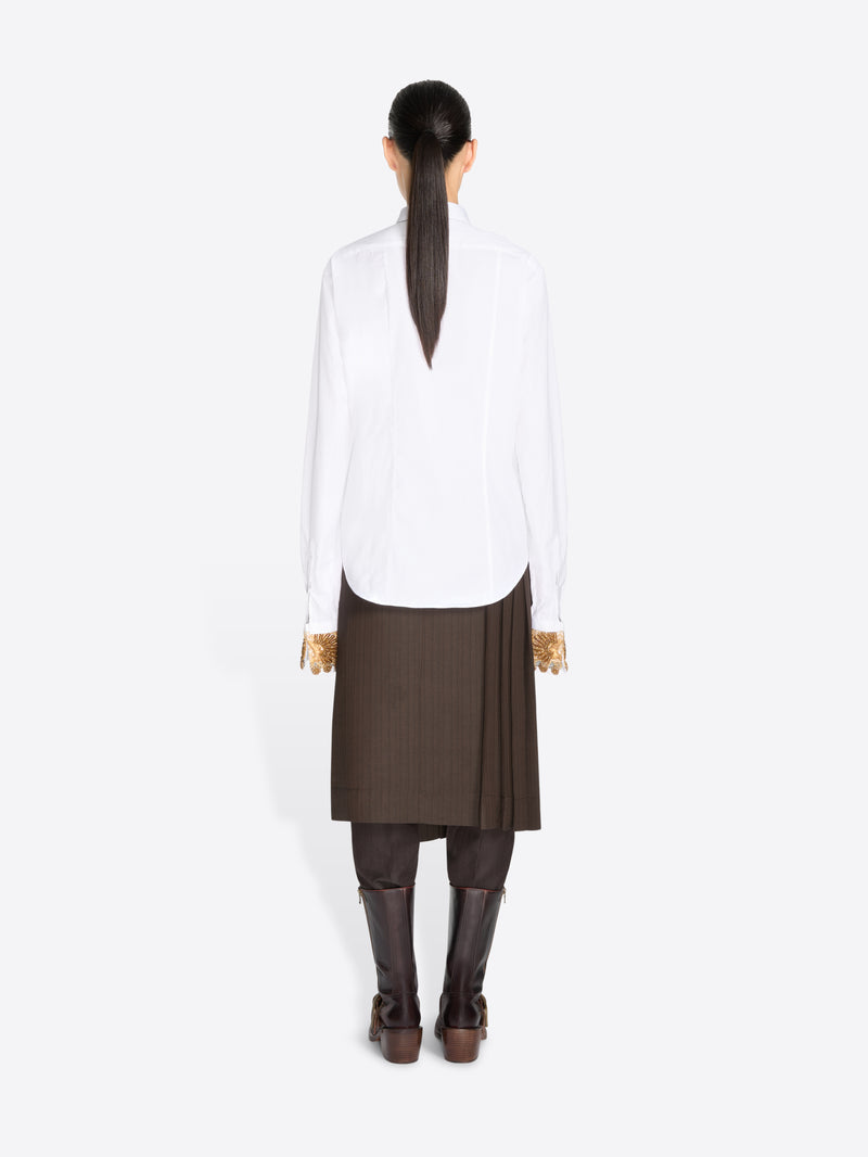 Back view of a model standing against a light gray background wearing a white long-sleeve button-up shirt, a brown pleated midi skirt, a brown tie, and dark brown knee-high boots.