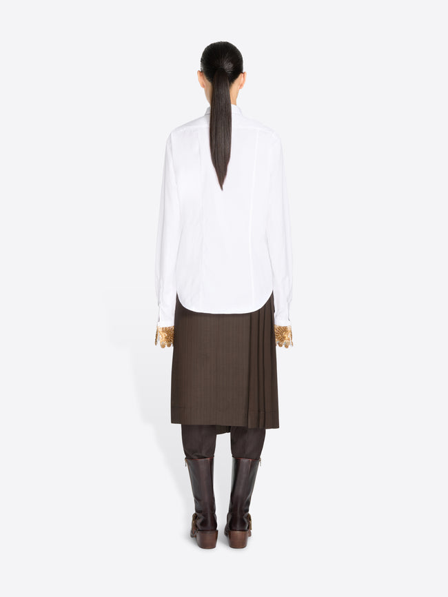 Back view of a model standing against a light gray background wearing a white long-sleeve button-up shirt, a brown pleated midi skirt, a brown tie, and dark brown knee-high boots.