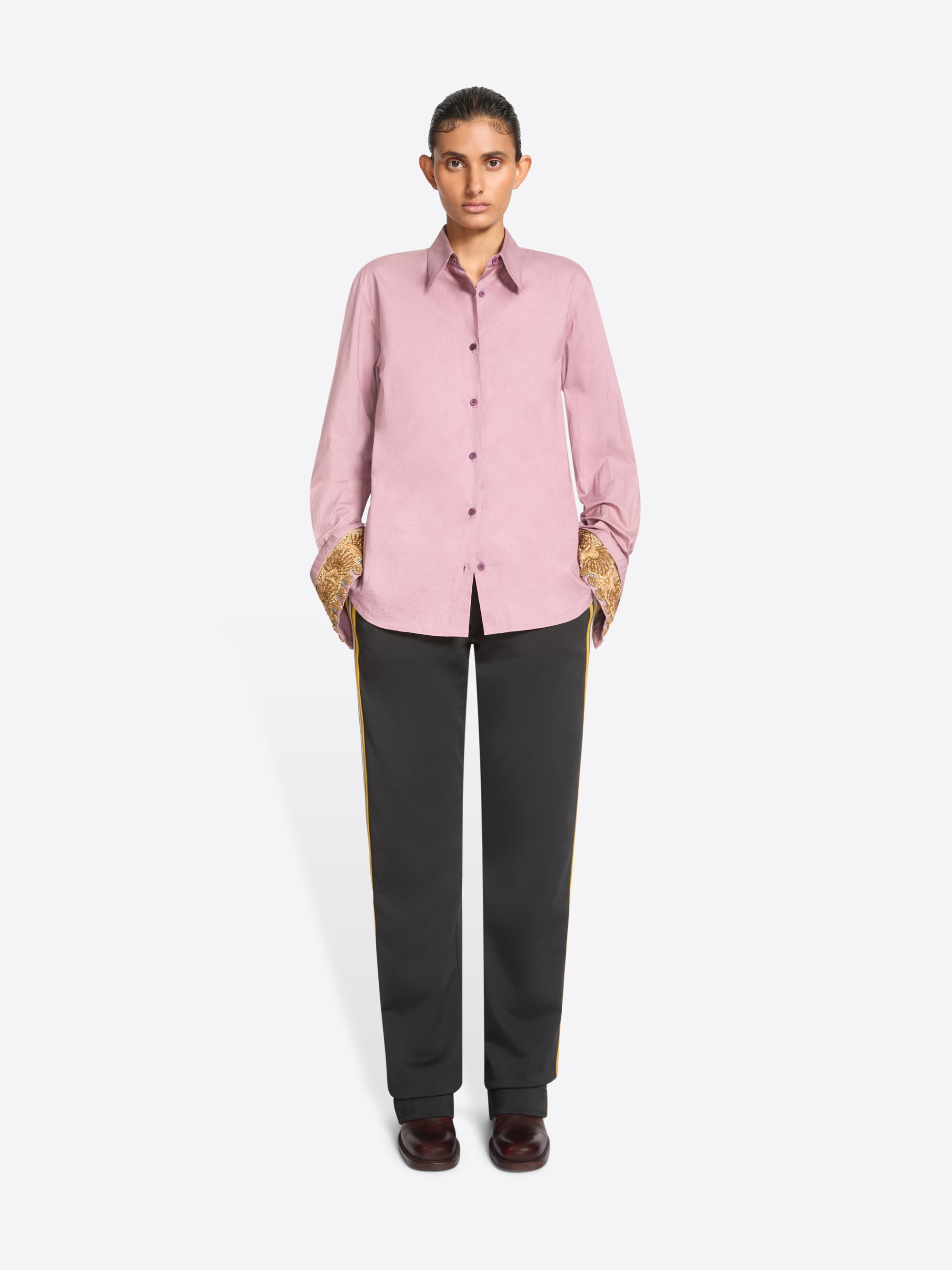 A model stands against a light gray background wearing a light pink long-sleeve button-up shirt with ornate gold patterned cuffs, paired with dark charcoal pants and dark brown shoes.
