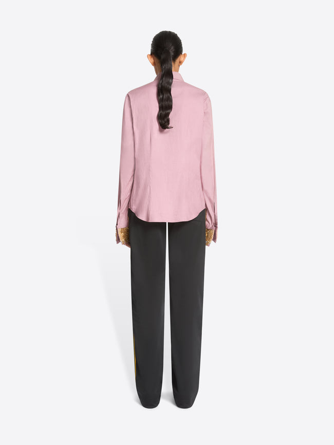 Back view of a model standing against a light gray background wearing a light pink long-sleeve button-up shirt with ornate gold patterned cuffs, paired with dark charcoal pants and dark brown shoes.