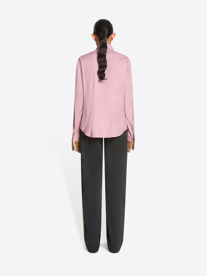 Back view of a model standing against a light gray background wearing a light pink long-sleeve button-up shirt with ornate gold patterned cuffs, paired with dark charcoal pants and dark brown shoes.
