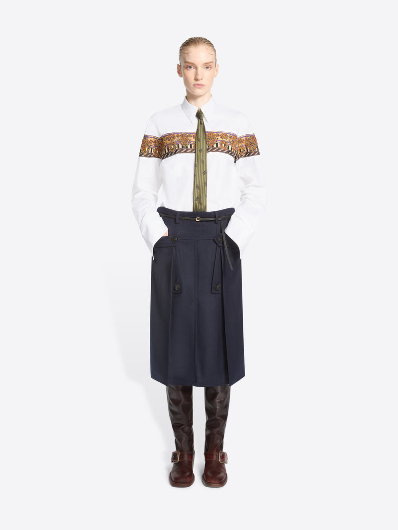 A model stands against a light gray background wearing a white long-sleeve shirt with a wide brown patterned band across the chest, a green patterned tie, a dark navy high-waisted belted skirt, and brown knee-high boots.