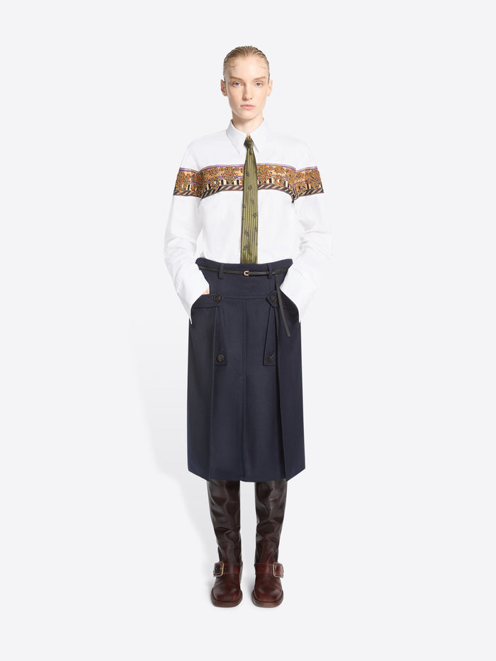 A model stands against a light gray background wearing a white long-sleeve shirt with a wide brown patterned band across the chest, a green patterned tie, a dark navy high-waisted belted skirt, and brown knee-high boots.
