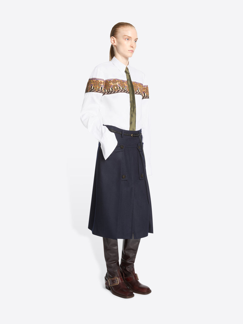 A model stands against a light gray background wearing a white long-sleeve shirt with a wide brown patterned band across the chest, a green patterned tie, a dark navy high-waisted belted skirt, and brown knee-high boots.