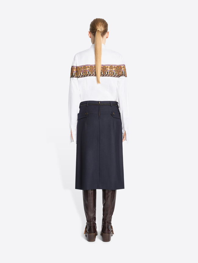 Back view of a model standing against a light gray background wearing a white long-sleeve shirt with a wide brown patterned band across the chest, a green patterned tie, a dark navy high-waisted belted skirt, and brown knee-high boots.