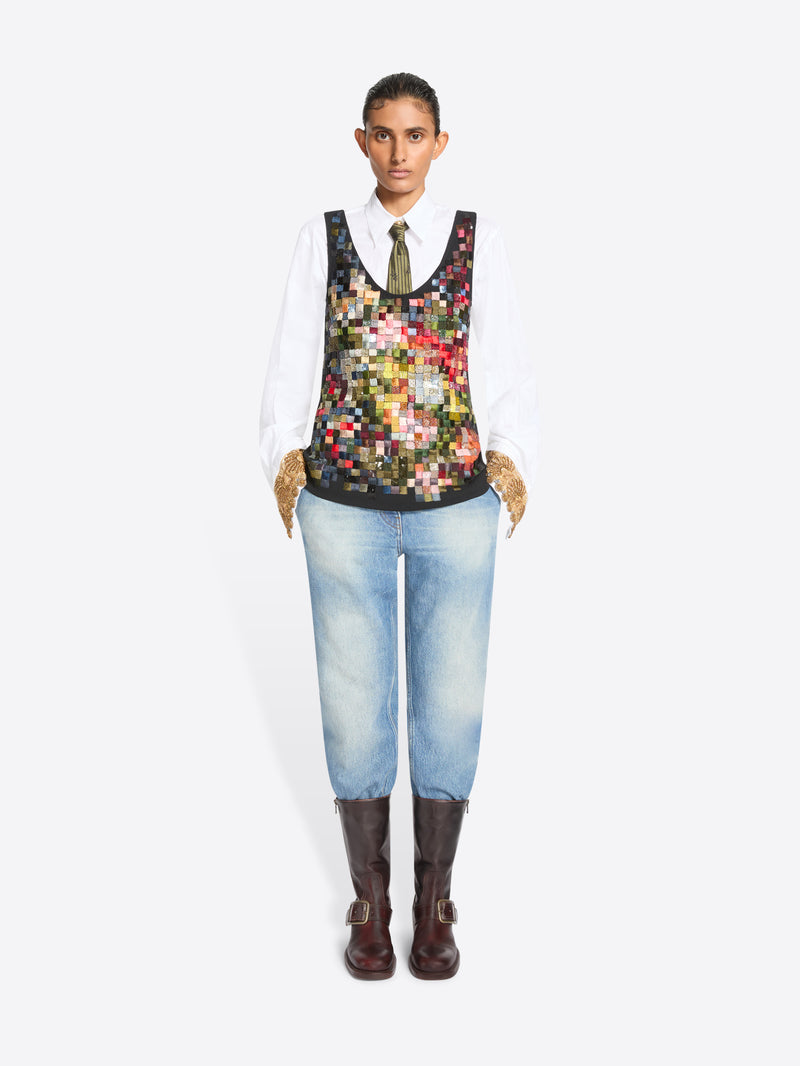 Full-length studio portrait of a person standing against a white background, wearing a crisp white shirt and patterned olive tie beneath a sleeveless multicolored patchwork vest, paired with light-wash jeans, textured tan gloves, and dark brown buckle boots.