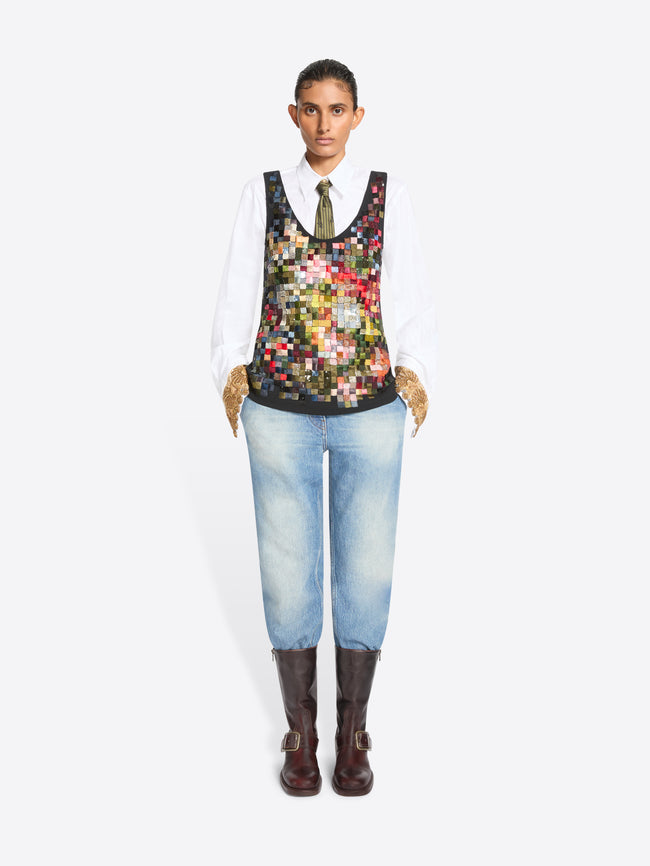Full-length studio portrait of a person standing against a white background, wearing a crisp white shirt and patterned olive tie beneath a sleeveless multicolored patchwork vest, paired with light-wash jeans, textured tan gloves, and dark brown buckle boots.