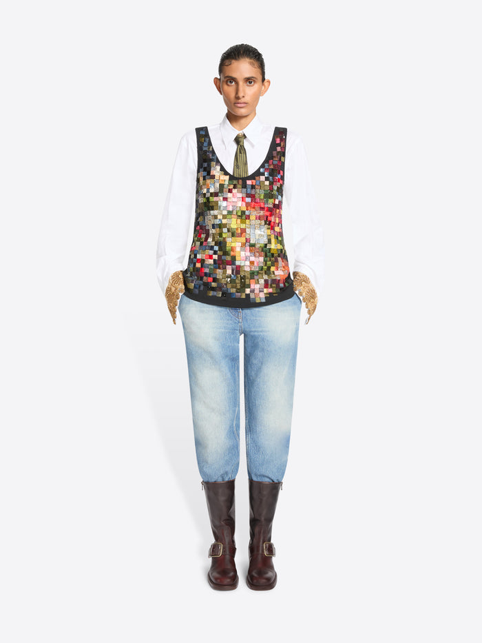 Full-length studio portrait of a person standing against a white background, wearing a crisp white shirt and patterned olive tie beneath a sleeveless multicolored patchwork vest, paired with light-wash jeans, textured tan gloves, and dark brown buckle boots.