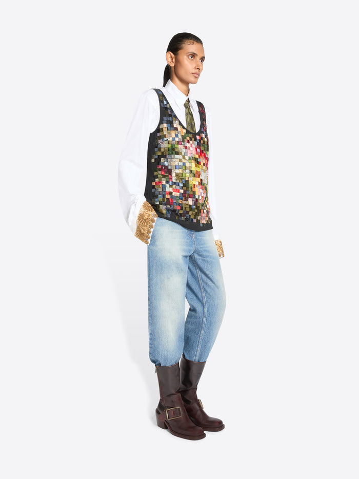 Full-length studio portrait of a person standing against a white background, wearing a crisp white shirt and patterned olive tie beneath a sleeveless multicolored patchwork vest, paired with light-wash jeans, textured tan gloves, and dark brown  buckle boots.