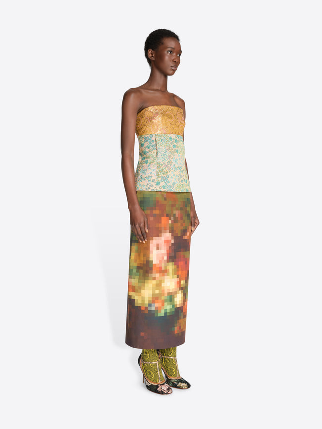 A model wearing a strapless top with a yellow-gold pattern and a multicolor abstract floral print skirt, paired with floral high heels, against a light gray background.