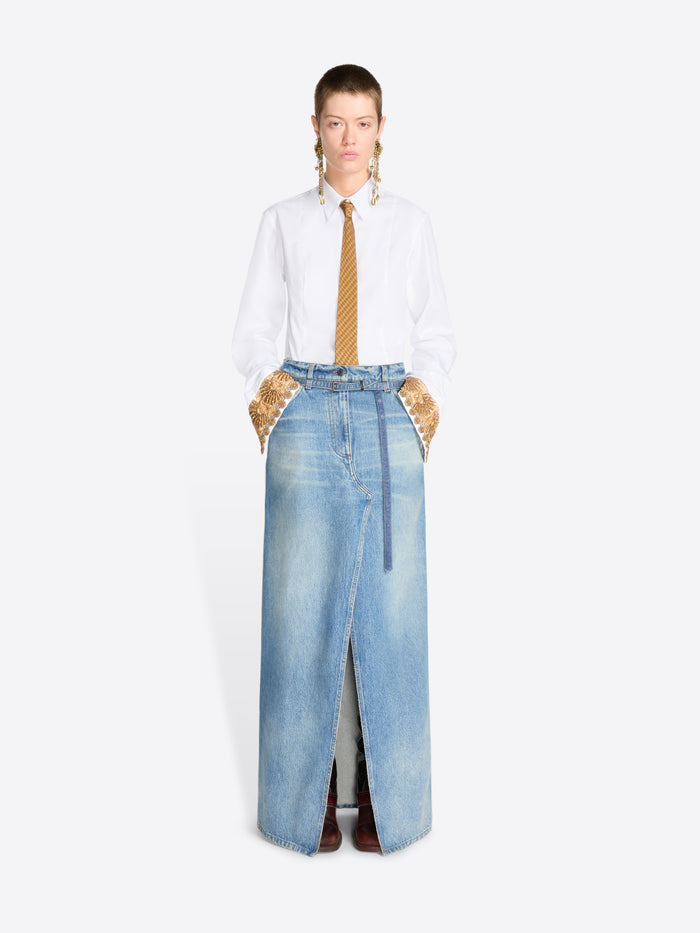 A model stands against a light gray background wearing a long light-wash denim skirt with a front slit, styled with a white button-up shirt and a gold tie.