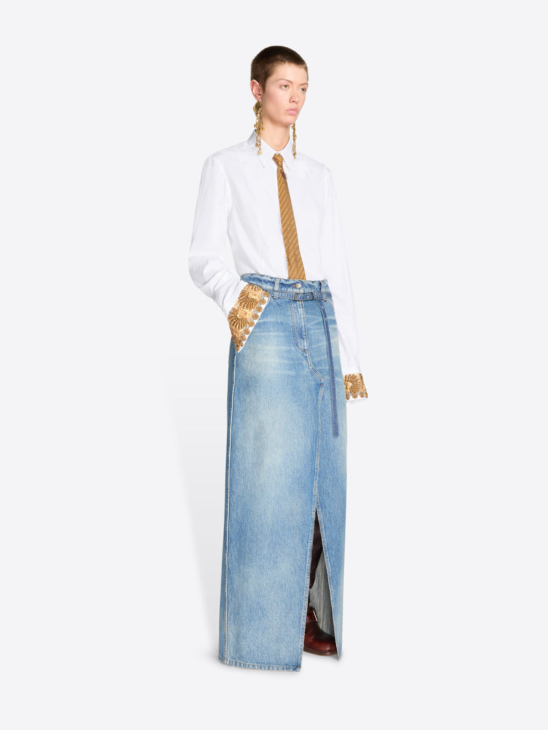 A model stands against a light gray background wearing a long light-wash denim skirt with a front slit, styled with a white button-up shirt and a gold tie.