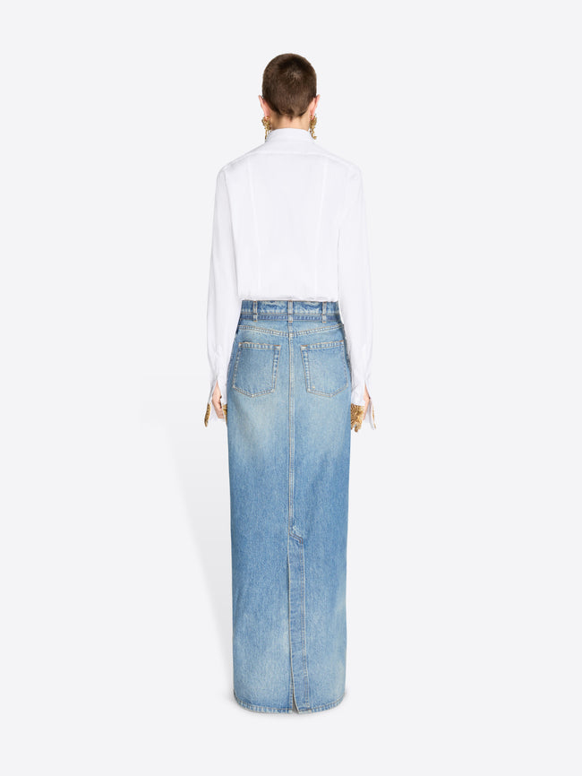 Back view of a model standing against a light gray background wearing a long light-wash denim skirt with a front slit, styled with a white button-up shirt.