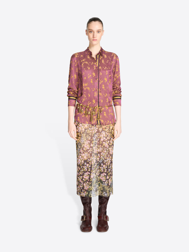 A model stands against a light gray background wearing a matching two-piece outfit consisting of a long-sleeve button-front shirt and a midi skirt, both in a dense floral and ornamental print.