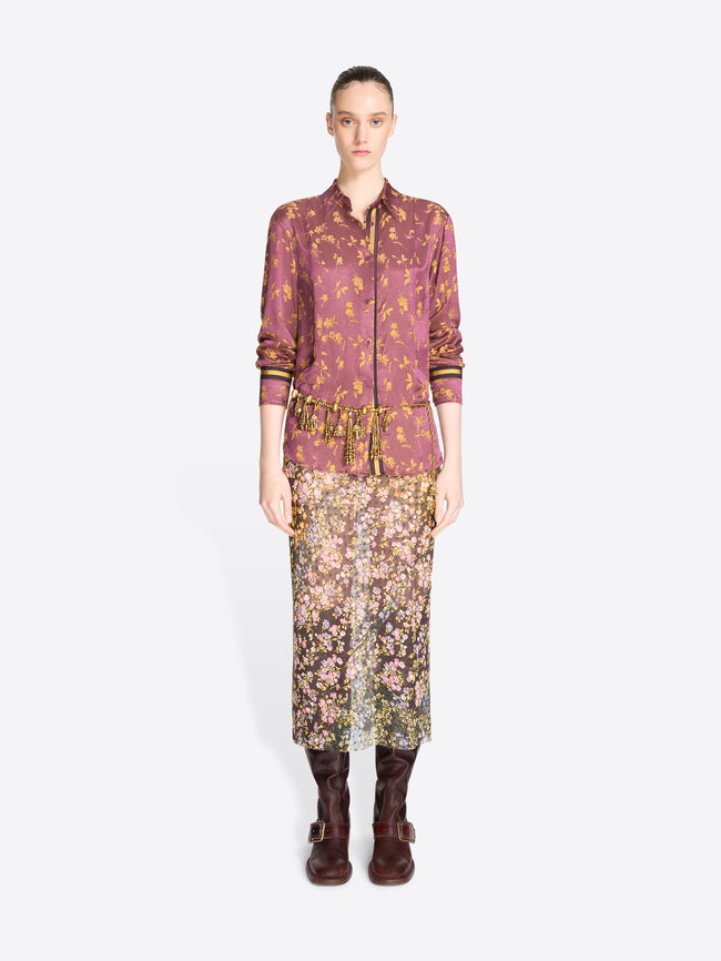 A model stands against a light gray background wearing a matching two-piece outfit consisting of a long-sleeve button-front shirt and a midi skirt, both in a dense floral and ornamental print.