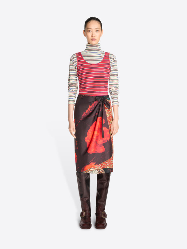 A model stands against a light gray background wearing a striped red tank top and gray striped turtleneck, a dark wrap skirt with a large red abstract print, and dark brown knee-high boots.
