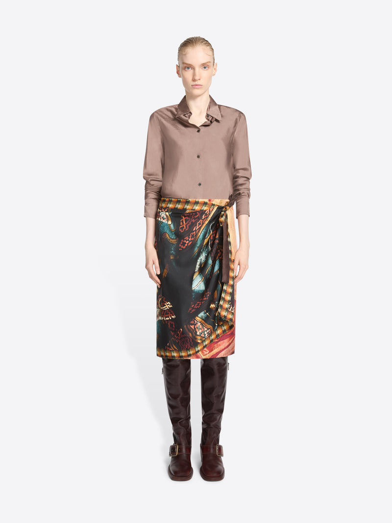 A model stands against a light gray background wearing a taupe button-up shirt, a dark wrap-style skirt with a colorful paisley and ornamental print, and dark brown knee-high boots.