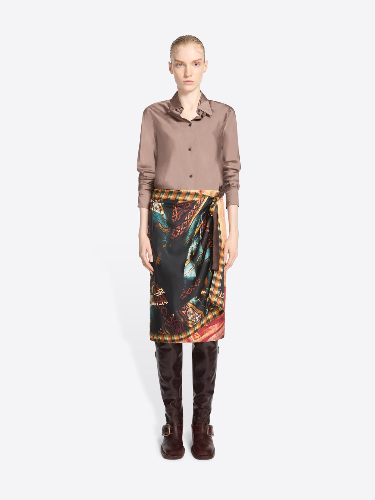A model stands against a light gray background wearing a taupe button-up shirt, a dark wrap-style skirt with a colorful paisley and ornamental print, and dark brown knee-high boots.