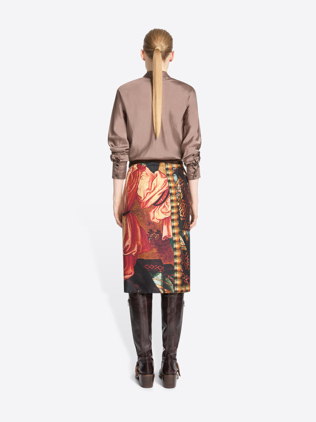 Back view of a model standing against a light gray background wearing a taupe button-up shirt, a dark wrap-style skirt with a colorful paisley and ornamental print, and dark brown knee-high boots.