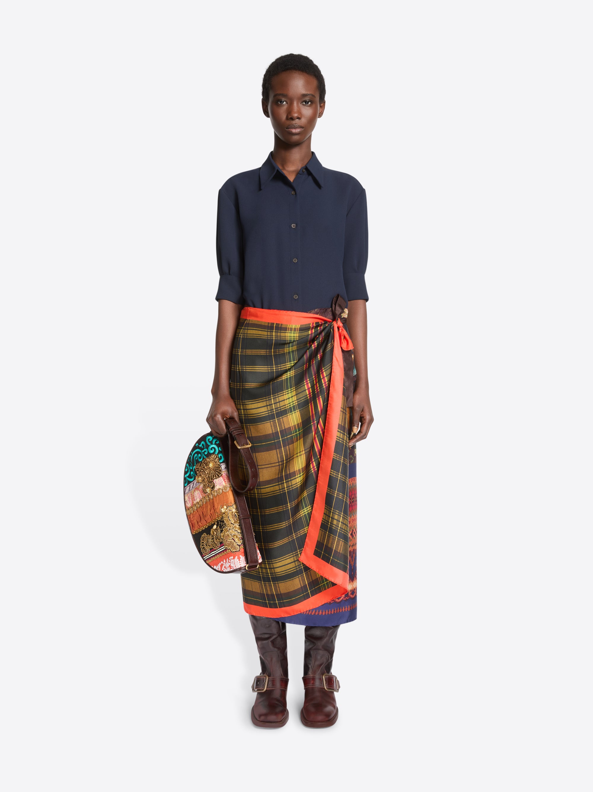 A model stands against a light gray background wearing a dark navy button-up shirt, a multicolor plaid wrap skirt with a bright orange border, and brown leather boots, holding a small round bag.