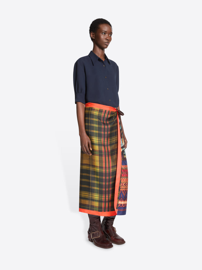 A model stands against a light gray background wearing a dark navy button-up shirt, a multicolor plaid wrap skirt with a bright orange border, and brown leather boots.