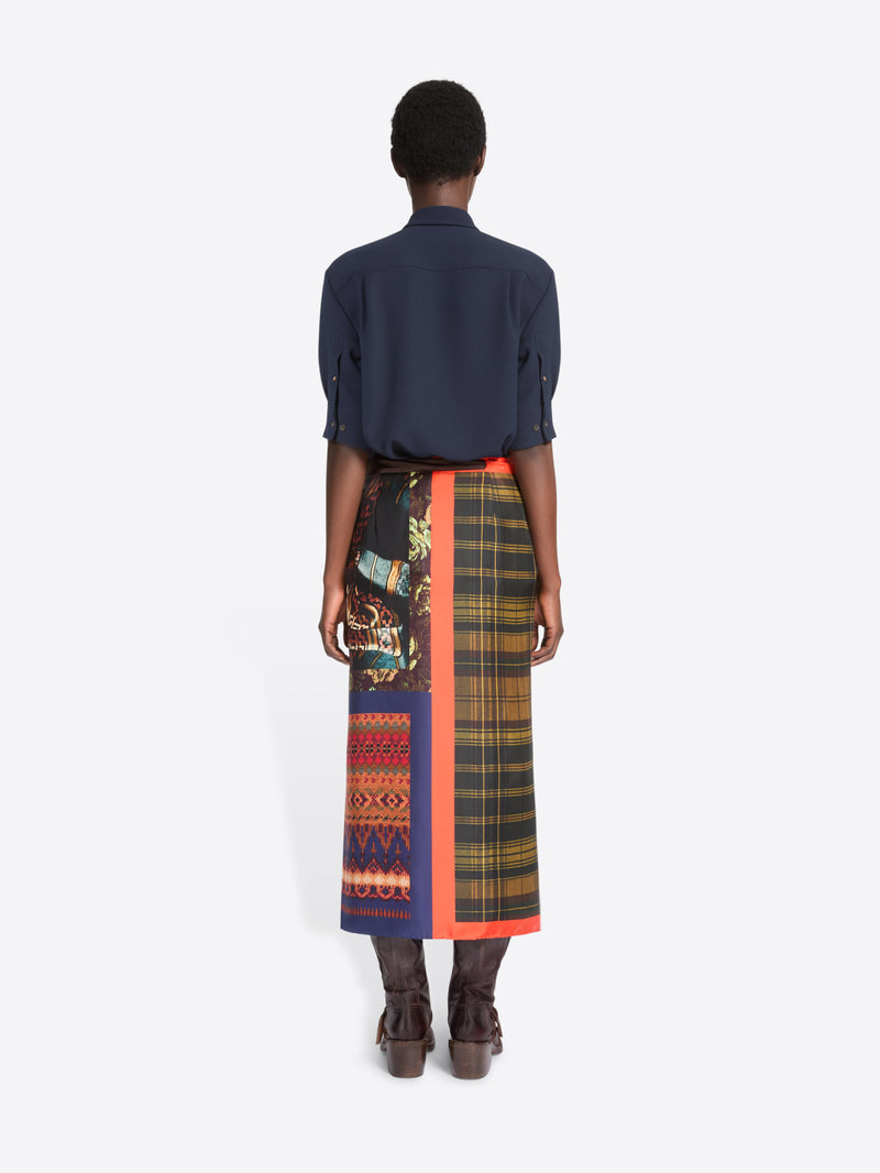 Back view of a model standing against a light gray background wearing a dark navy button-up shirt, a multicolor plaid wrap skirt with a bright orange border, and brown leather boots.