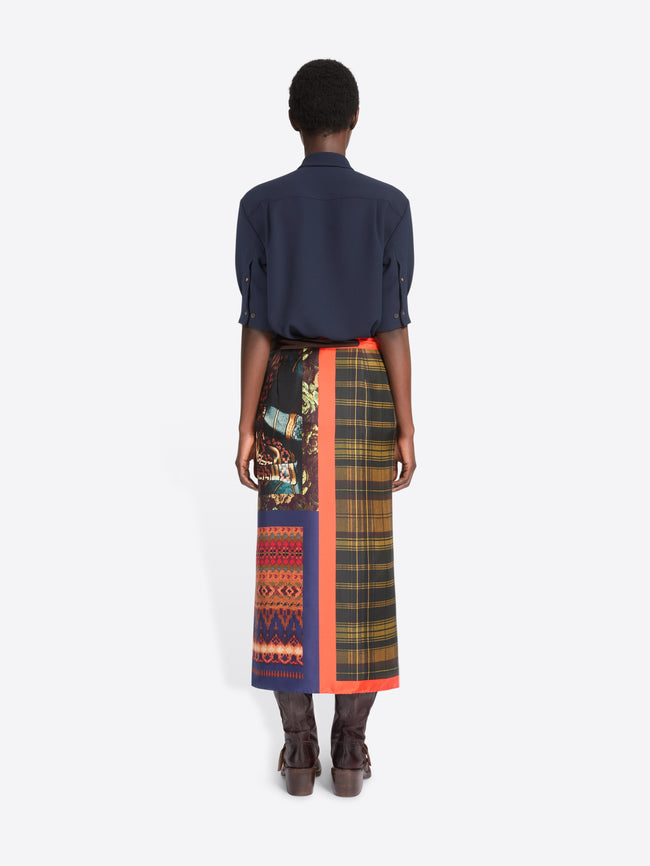 Back view of a model standing against a light gray background wearing a dark navy button-up shirt, a multicolor plaid wrap skirt with a bright orange border, and brown leather boots.