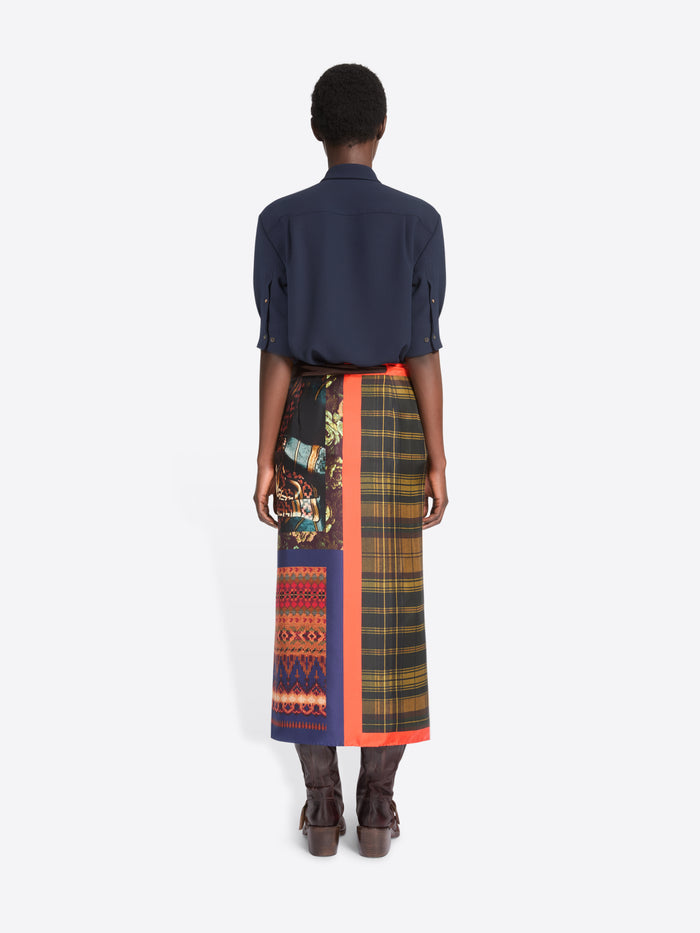 Back view of a model standing against a light gray background wearing a dark navy button-up shirt, a multicolor plaid wrap skirt with a bright orange border, and brown leather boots.