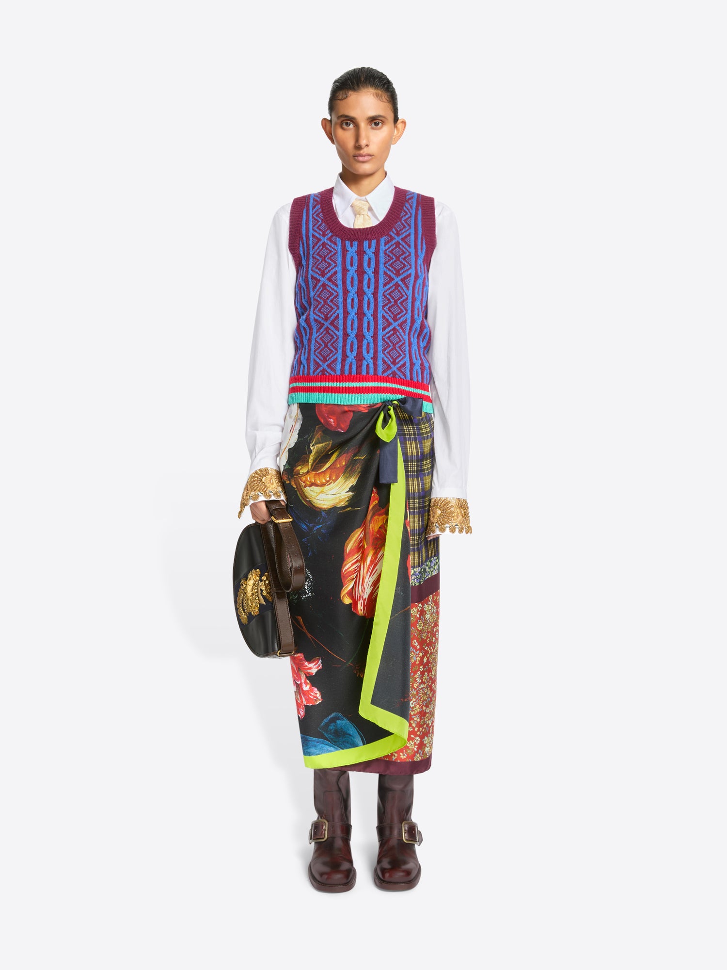A model stands against a light gray background wearing a white long-sleeve shirt, a colorful patterned knit sweater vest, a long multicolor skirt and dark brown leather boots, holding a black bag with gold detailing.