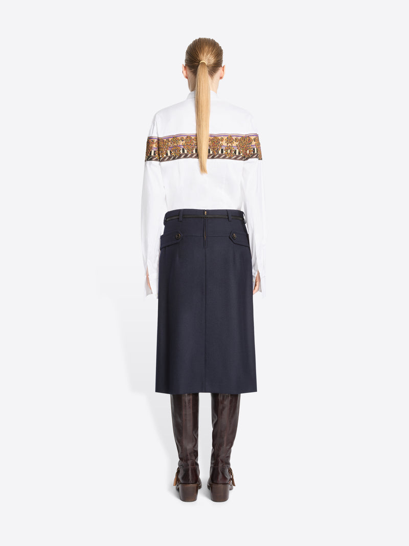 Back view of model standing against a light gray background wearing a white long-sleeve shirt with a wide brown patterned band across the chest, a green patterned tie, a dark navy high-waisted belted skirt, and brown knee-high boots.
