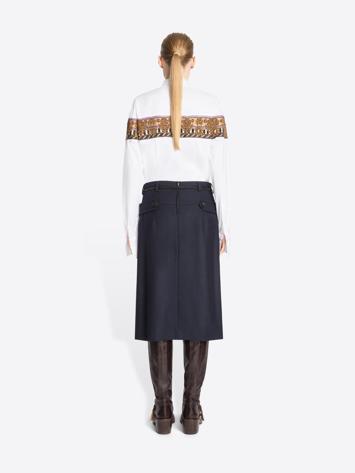 Back view of model standing against a light gray background wearing a white long-sleeve shirt with a wide brown patterned band across the chest, a green patterned tie, a dark navy high-waisted belted skirt, and brown knee-high boots.