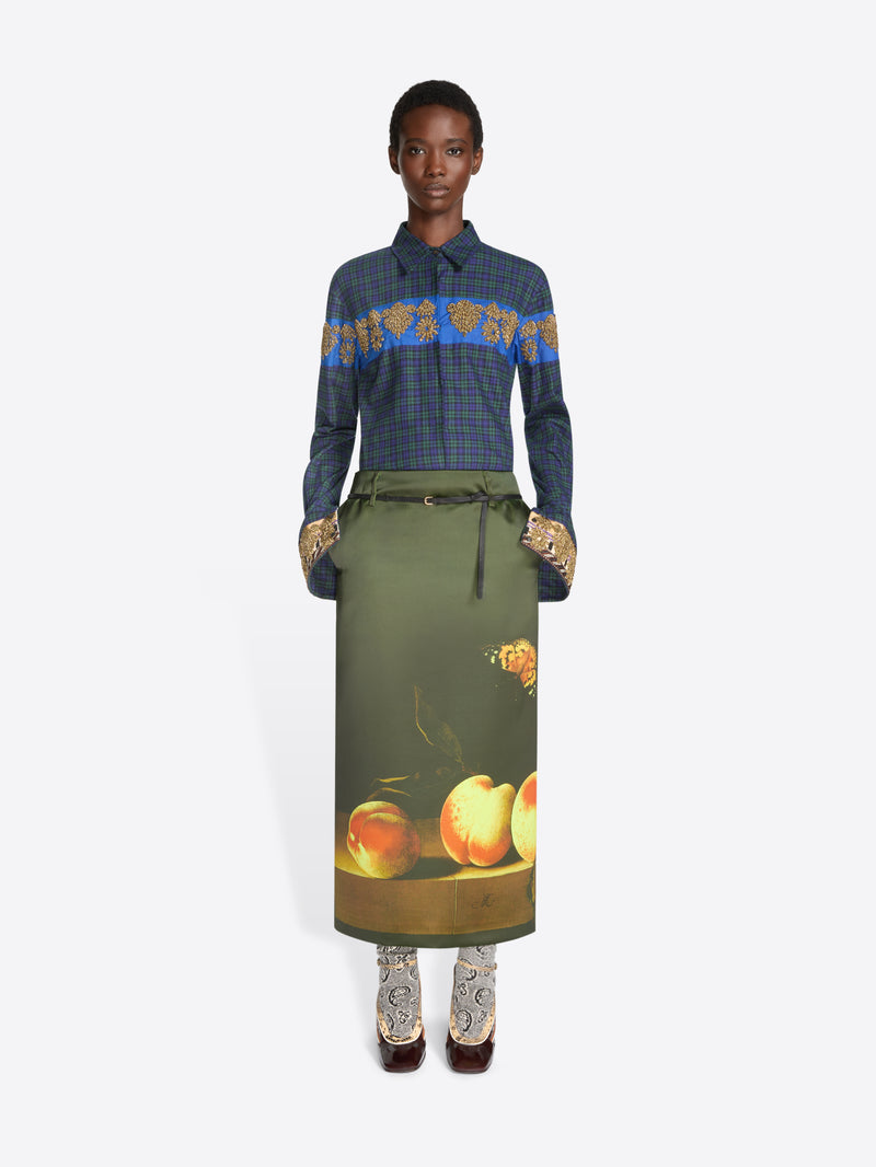 A model stands against a light gray background wearing a long-sleeve plaid shirt with a blue and gold patterned yoke, an olive skirt with a fruit print near the hem and pointed heels.