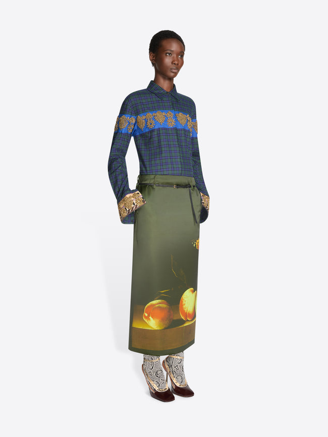 A model stands against a light gray background wearing a long-sleeve plaid shirt with a blue and gold patterned yoke, an olive skirt with a fruit print near the hem and pointed heels.