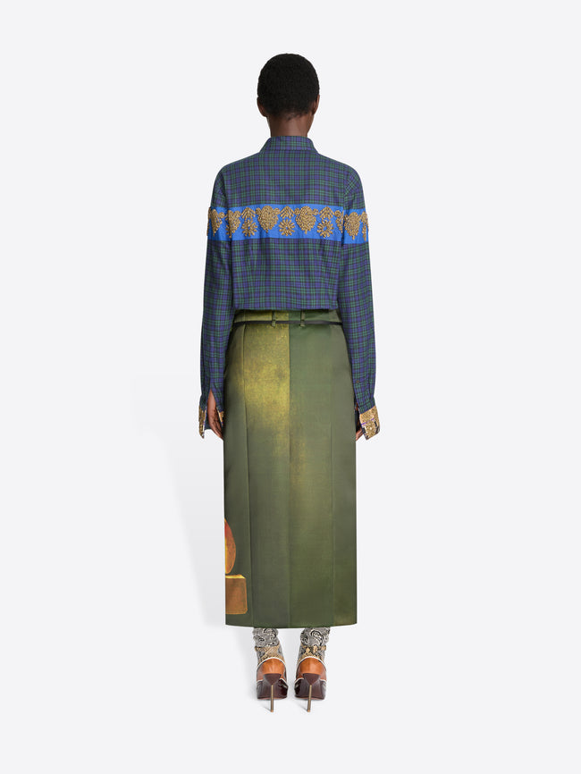 Back view of a model standing against a light gray background wearing a long-sleeve plaid shirt with a blue and gold patterned yoke, an olive skirt with a fruit print near the hem and pointed heels.