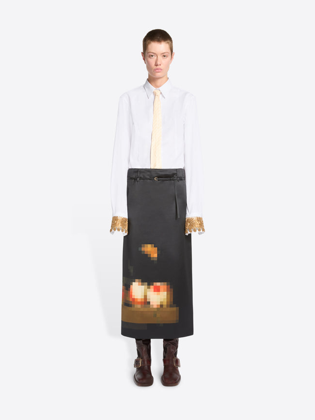 A model stands against a light gray background wearing a white long-sleeve shirt with gold lace cuffs, a cream tie, and a black skirt featuring a pixel-art flame graphic near the hem, paired with dark brown boots.